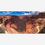 Panoramic photo of a meandering canyon with red rock formations under a blue sky with clouds.