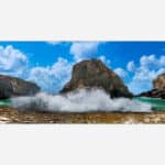 A panoramic photo of ocean waves crashing against rocky sea cliffs under a blue sky with fluffy clouds.