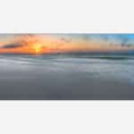 A panoramic photo of a serene beach at sunset with vibrant colors in the sky and gentle waves in the foreground.