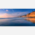A serene coastal landscape at sunset with reflective ocean waters and a striking cliff formation on the right.