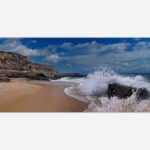 Image shows a dynamic beach scene with a wave crashing against a rock on the sandy shore with a clear blue sky and coastal cliffs in the background.