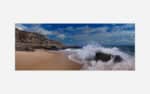 Image shows a dynamic beach scene with a wave crashing against a rock on the sandy shore with a clear blue sky and coastal cliffs in the background.