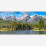 A panoramic photograph of a mountain landscape with a forest and lake in the foreground and a blue sky above.