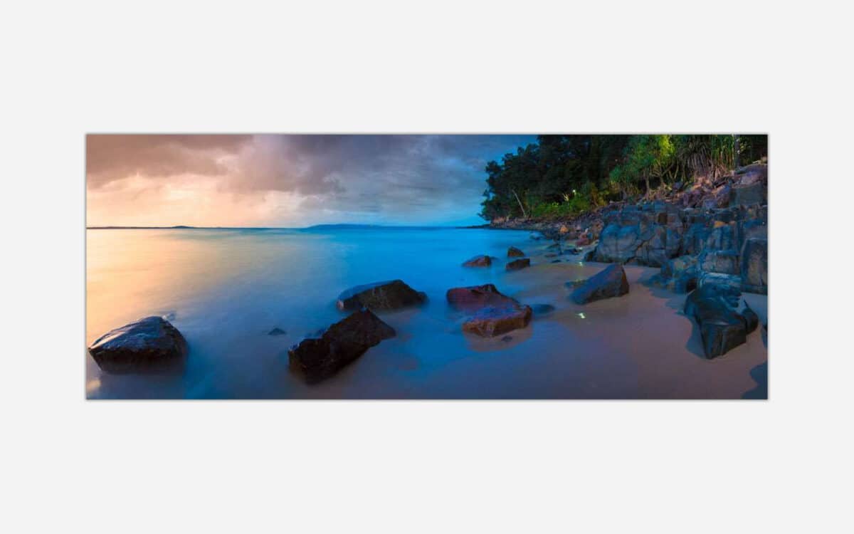 Alt text: A serene seascape photograph capturing a beach at sunset with smooth water, a rocky shoreline, and a twilight sky with soft blue and orange hues.