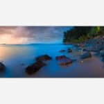 Alt text: A serene seascape photograph capturing a beach at sunset with smooth water, a rocky shoreline, and a twilight sky with soft blue and orange hues.