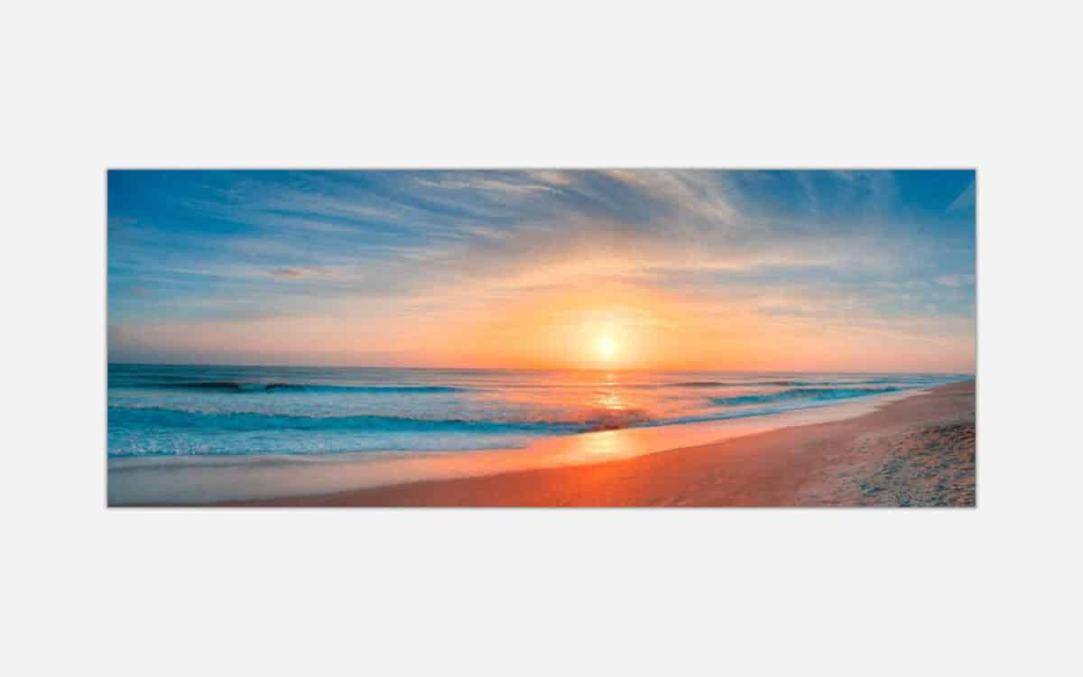 A panoramic view of a beach at sunset with the sun reflecting on the water and wet sand, featuring a colorful sky and gentle waves.