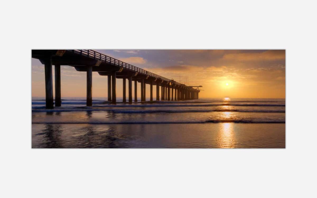 A photograph of a pier extending over the ocean with a stunning sunset in the background, casting warm golden light across the scene.