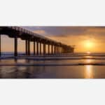 A photograph of a pier extending over the ocean with a stunning sunset in the background, casting warm golden light across the scene.