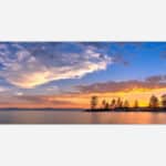 A panoramic photograph of a sunset with golden and orange hues in the sky, silhouetted trees against a calm lake with dramatic clouds above.
