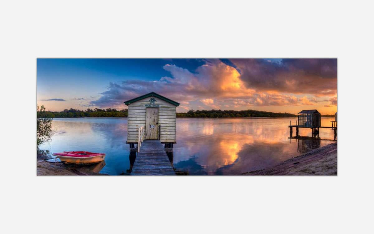 A panoramic art print of a serene lake scene at sunset, featuring a wooden boat dock with a small boat, calm water reflecting the colorful sky, and a quaint boat house at the end of the pier.