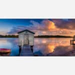 A panoramic art print of a serene lake scene at sunset, featuring a wooden boat dock with a small boat, calm water reflecting the colorful sky, and a quaint boat house at the end of the pier.