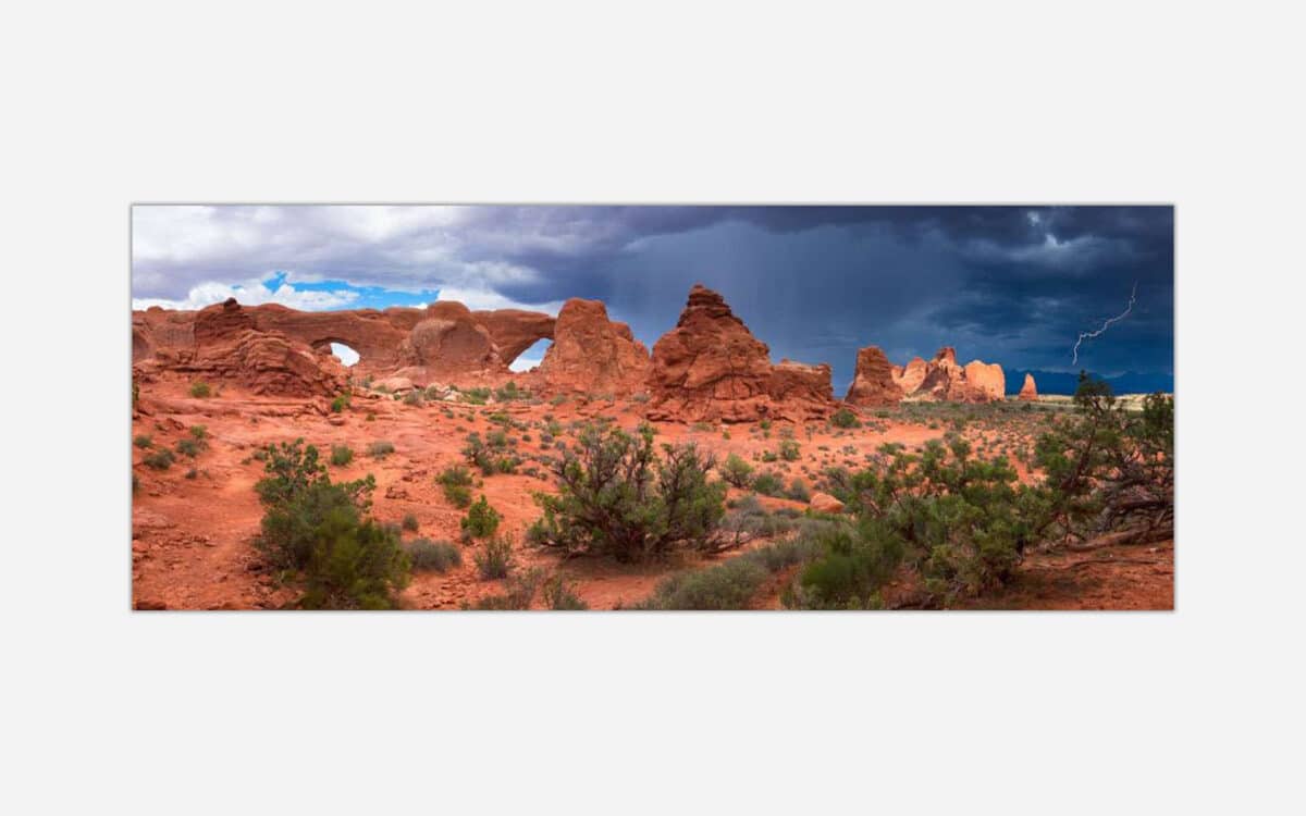 A panoramic photograph of Arches National Park with dramatic storm clouds and a lightning bolt in the distance over red desert rock formations.