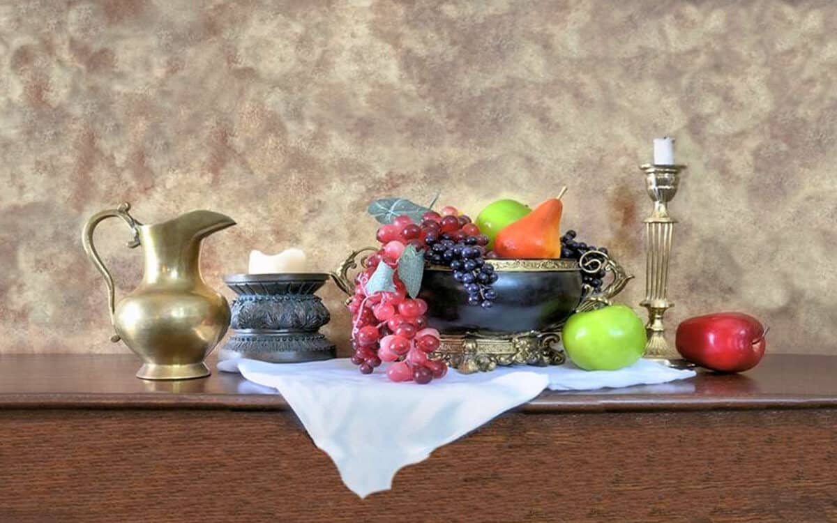 A still life painting featuring a brass pitcher, fruit bowl with grapes, pears, and apples, a burning candle in a holder, and antique items on a draped white cloth with a textured background.
