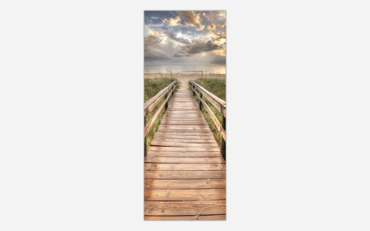 A vertical photograph of a wooden boardwalk leading through beach grass towards the ocean with a cloudy sky at sunset.
