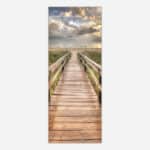 A vertical photograph of a wooden boardwalk leading through beach grass towards the ocean with a cloudy sky at sunset.